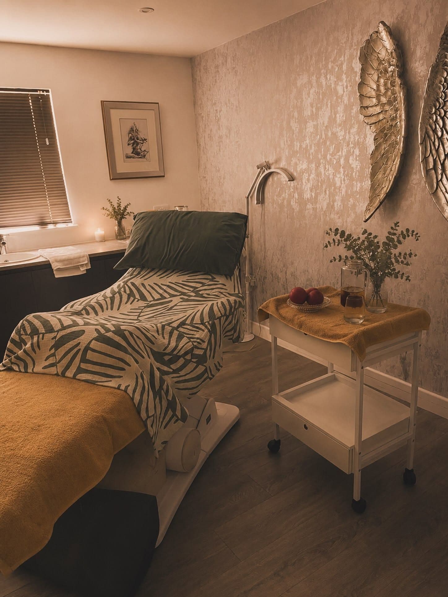 Nurture Body Therapy treatment room — palm-print bedding, warm candle light, silver angel-wing wall art. Ashford, Kent.