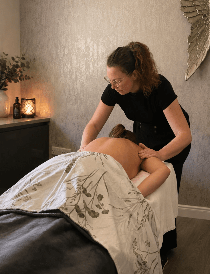 Kim Homewood at work — hands resting gently on a client's back during a treatment at Nurture Body Therapy in Ashford, Kent. Calm, low-lit treatment room with eucalyptus, candles and angel-wing wall art.