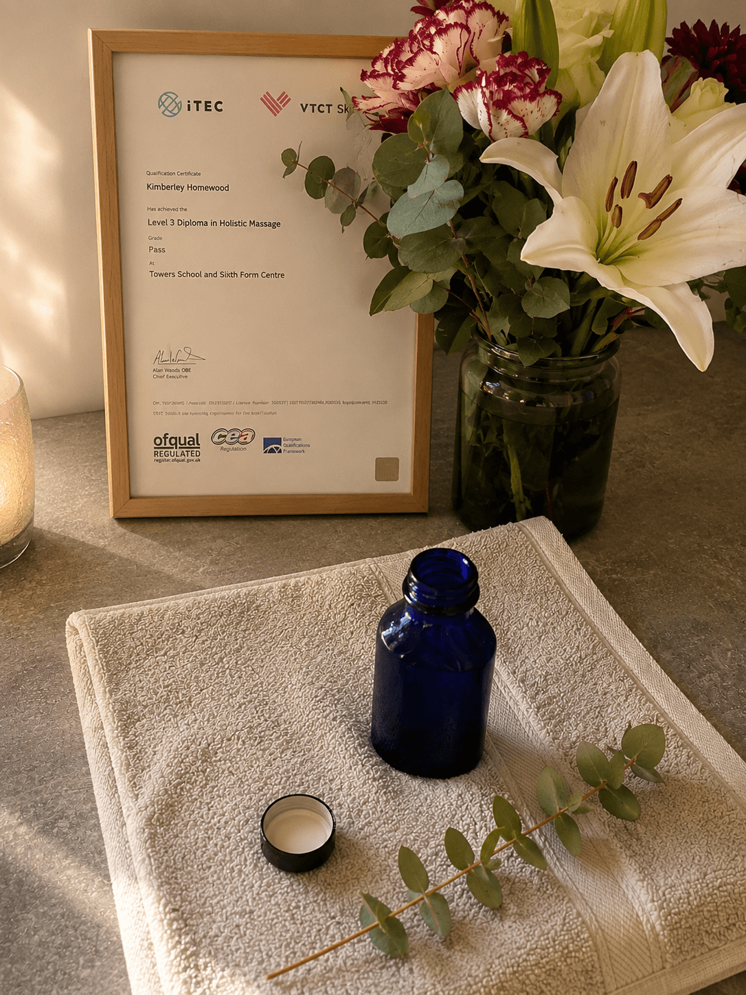 Kim Homewood's iTEC Level 3 Diploma in Holistic Massage certificate displayed beside a Neal's Yard Remedies apothecary bottle, eucalyptus and fresh flowers — Nurture Body Therapy treatment room